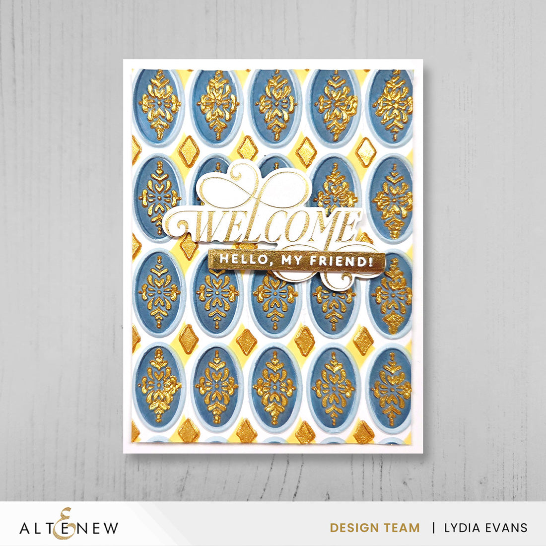 A handmade greeting card with a white border, featuring a repeating pattern of blue and gold ornate medallions and the text 'WELCOME HELLO, MY FRIEND!' in layered white and gold lettering. The card is photographed against a light gray wood-textured background, with a white banner at the bottom containing the brand name ALTENEW and designer credit.