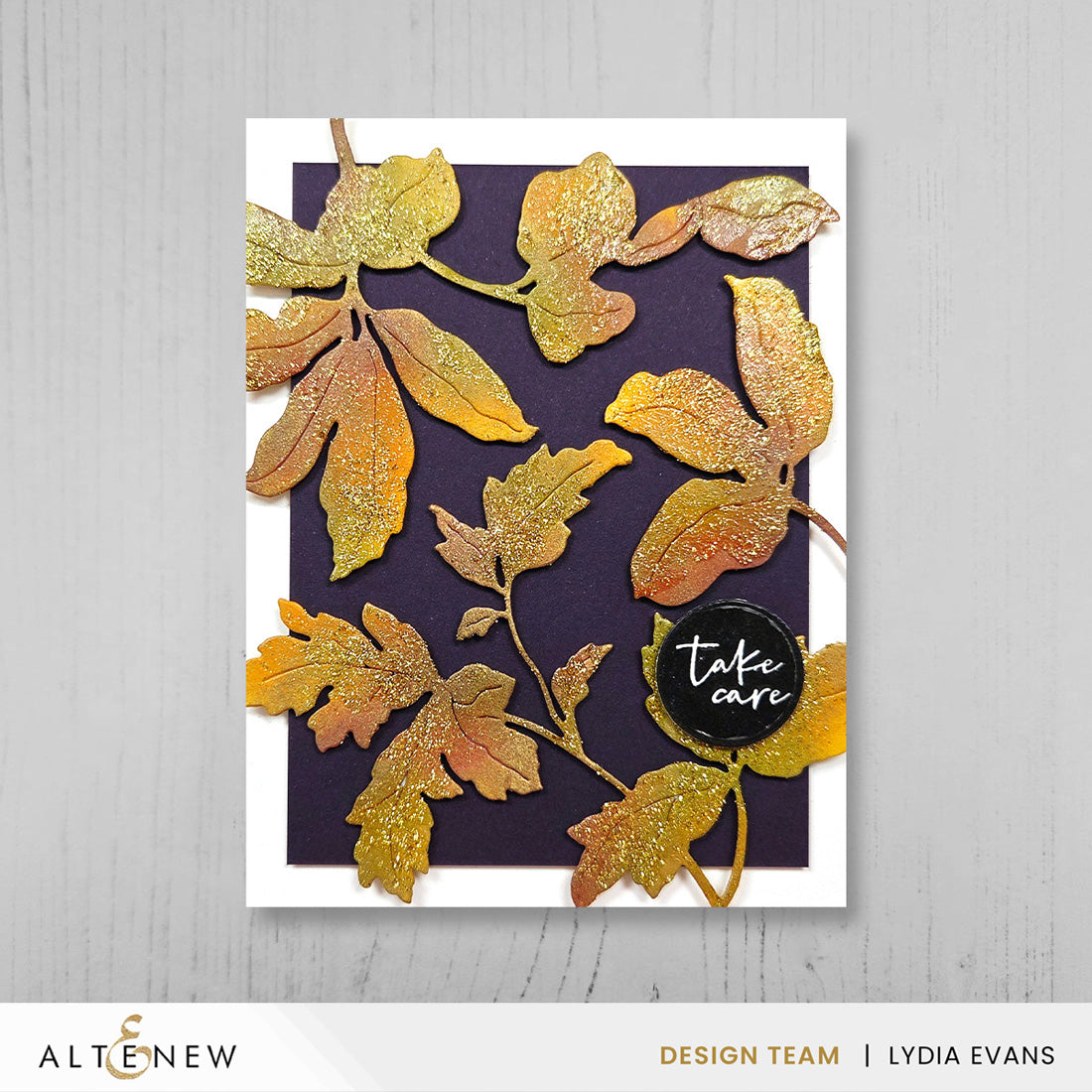 A handmade greeting card featuring layered glittery gold and orange autumn leaves on a dark purple background, with a small black circular badge that reads 'take care'. The card is displayed on a light gray wood-textured background, with the Altenew and Lydia Evans branding visible at the bottom.