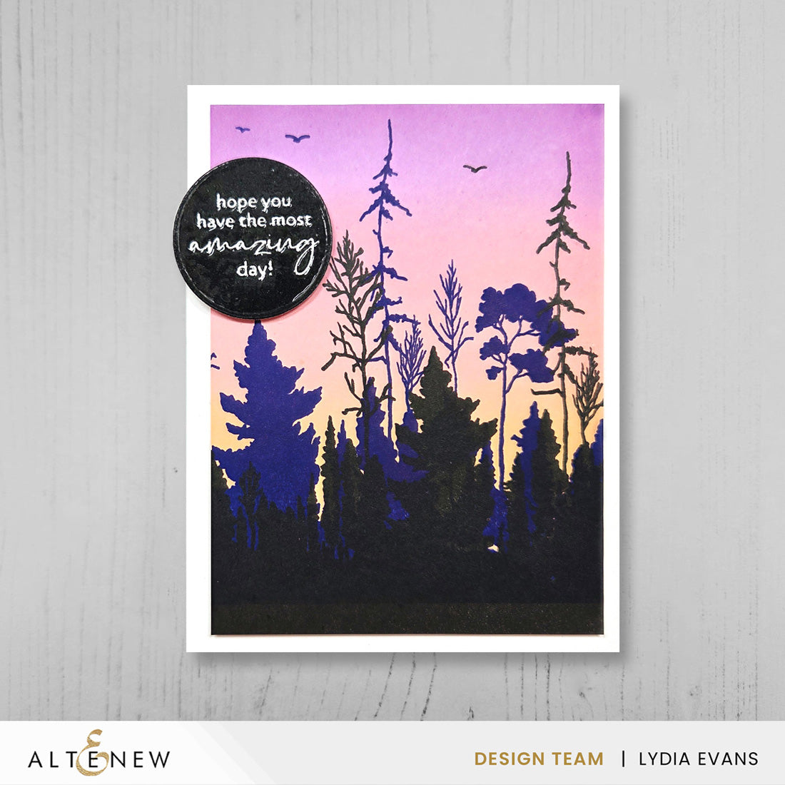 A handmade greeting card featuring a forest silhouette against a pink, purple, and orange gradient sky, with a black circular sticker that reads 'hope you have the most amazing day!'. The card is displayed on a light gray wood-textured background.