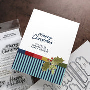 A handmade Christmas greeting card with navy blue script and block text, a striped blue, red, and yellow band, and a holly graphic, displayed with clear stamps and metal dies on a brown textured surface.
