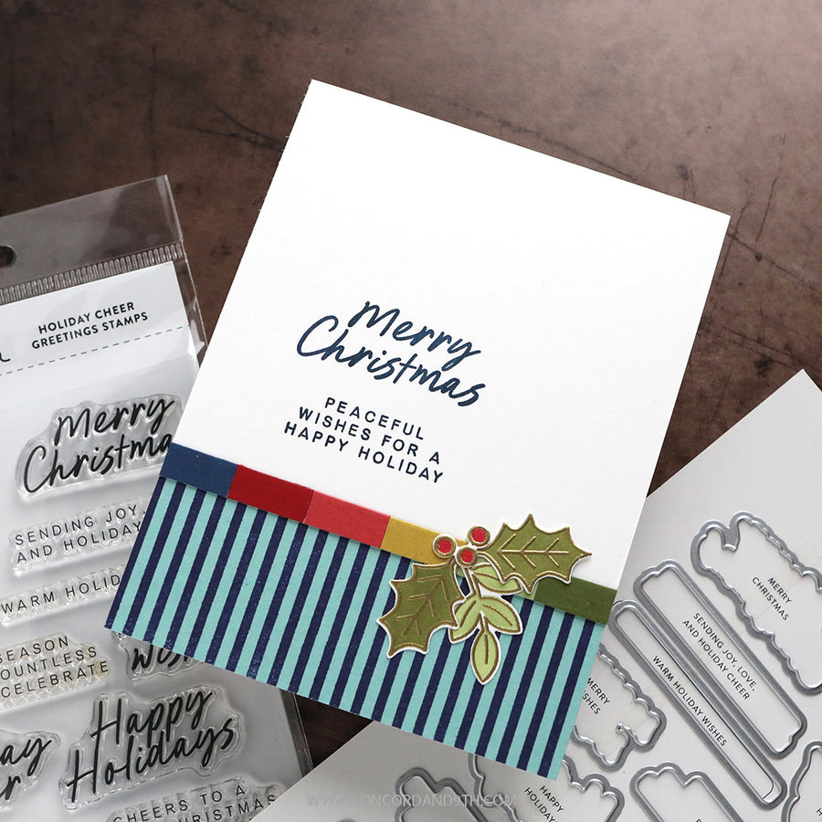 A handmade Christmas greeting card with navy blue script and block text, a striped blue, red, and yellow band, and a holly graphic, displayed with clear stamps and metal dies on a brown textured surface.