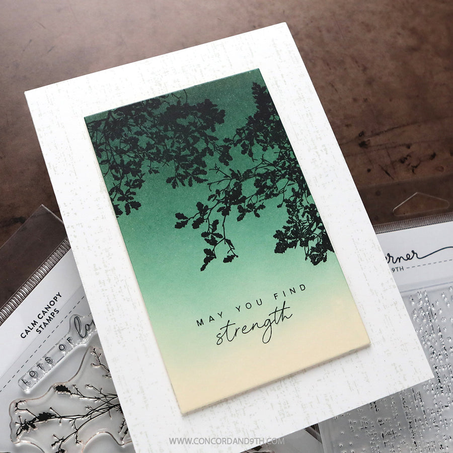 A handmade greeting card with a white textured border and a central printed panel featuring a green-to-cream gradient, black leafy branch silhouette, and the words 'MAY YOU FIND strength.' The card is photographed on a brown surface with clear stamp blocks partially visible around it.
