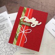 A handmade red Christmas greeting card with a green and red striped ribbon, a white bow, and a 'Merry & Bright' die-cut embellishment, displayed on a tabletop with crafting supplies.