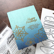 A handmade holiday greeting card with a blue gradient background, gold foil snowflakes and Christmas tree outlines, and gold foil 'Happy Holidays' text, displayed on a wooden surface with stamp and die craft supplies around it.