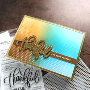 A handmade greeting card with a gold and blue gradient background, featuring a raised metallic gold 'Thankful' die-cut element and a small gold banner reading 'FOR YOUR PRAYERS'. The card is styled on top of clear stamp sheets on a textured brown surface.