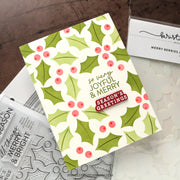 A handmade Christmas greeting card with a green and white holly berry pattern, displayed on a dark wood surface with crafting tools and stamps.