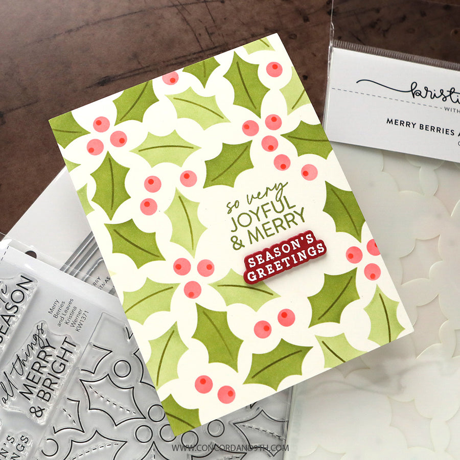 A handmade Christmas greeting card with a green and white holly berry pattern, displayed on a dark wood surface with crafting tools and stamps.