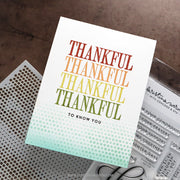 A handmade greeting card with the word 'THANKFUL' repeated in a vertical gradient of red, orange, yellow, and green, fading into a light blue-green dotted pattern at the bottom. The card is styled on a dark brown textured surface next to a metal perforated sheet and a clear stamp block.