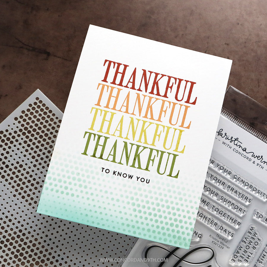 A handmade greeting card with the word 'THANKFUL' repeated in a vertical gradient of red, orange, yellow, and green, fading into a light blue-green dotted pattern at the bottom. The card is styled on a dark brown textured surface next to a metal perforated sheet and a clear stamp block.