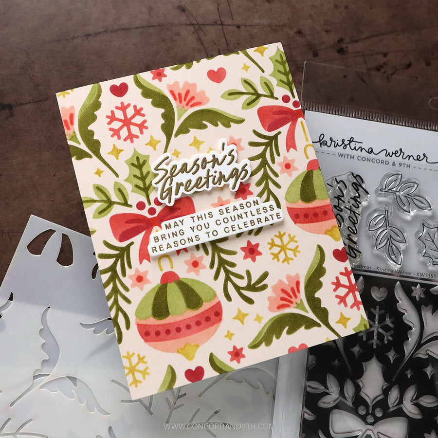 A handmade holiday greeting card with a festive pattern of evergreen branches, snowflakes, and ornaments, displayed alongside crafting tools including a clear stamp block and metal dies on a dark wooden surface.