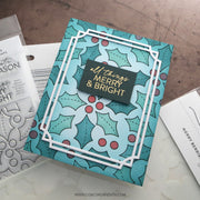 A handmade Christmas greeting card featuring a teal and white holly pattern with red berries and a central black label reading 'all things MERRY & BRIGHT' in gold foil, styled on a white surface with crafting tools partially visible.