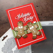 A handmade Christmas greeting card with two gingerbread men, shown on a rustic brown background with crafting tools partially visible underneath.
