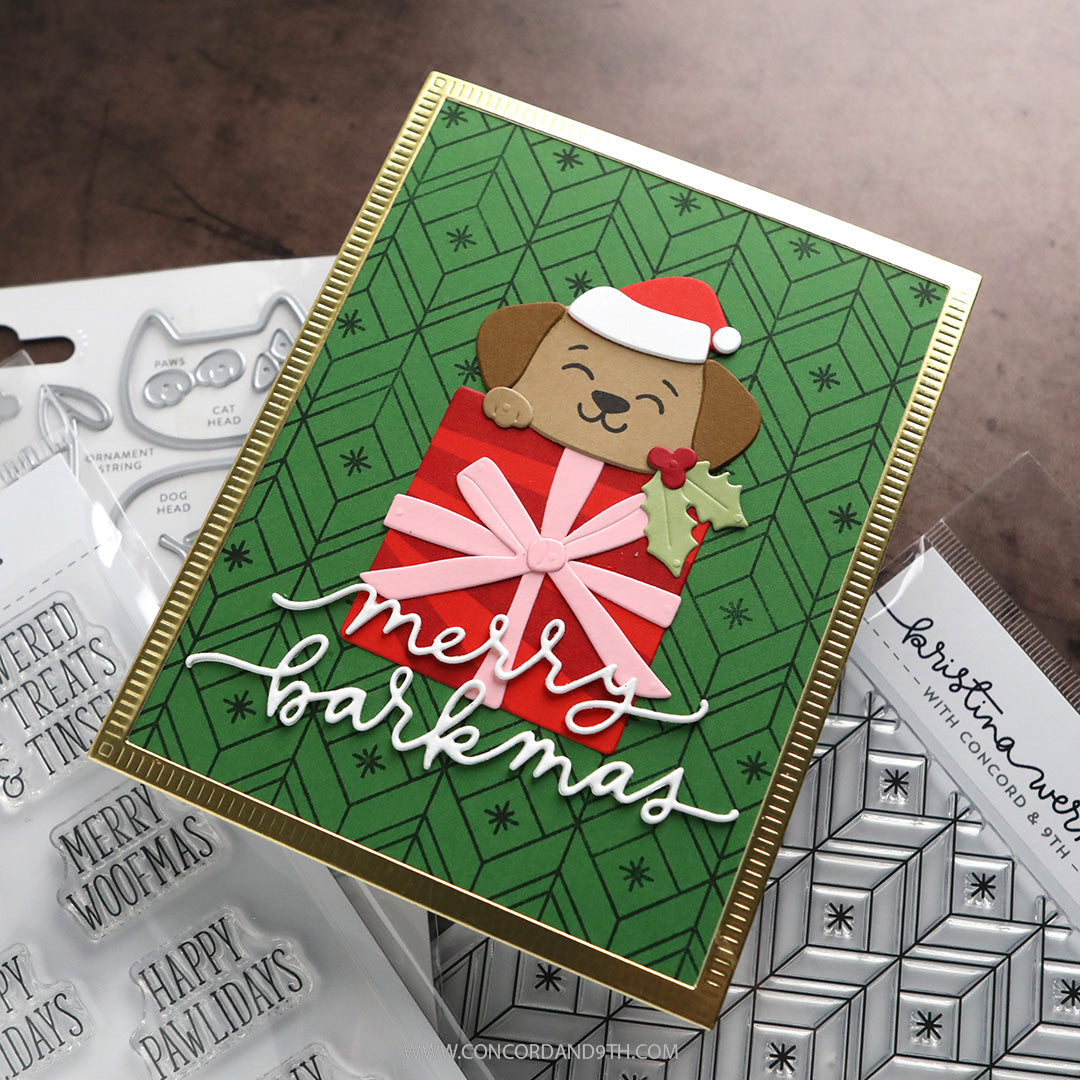 A handmade Christmas greeting card featuring a brown dog wearing a Santa hat and a red gift bag with a pink bow, set against a green patterned background with white script reading 'merry barkmas'. The card is styled on top of crafting supplies, including clear stamps and patterned sheets.