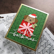 A handmade Christmas greeting card featuring a brown dog wearing a Santa hat and a red gift bag with a pink bow, set against a green patterned background with white script reading 'merry barkmas'. The card is styled on top of crafting supplies, including clear stamps and patterned sheets.