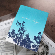A handmade greeting card with a blue gradient background, navy blue floral silhouette at the bottom, and the words 'HEARTFELT hugs' in white script, displayed on top of craft supplies.