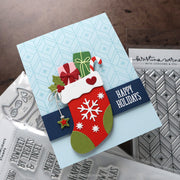 A handmade Christmas greeting card with a red stocking, green gift box, candy cane, holly, and a navy banner reading “HAPPY HOLIDAYS,” photographed on a tabletop with stamp and embosser tools around it.