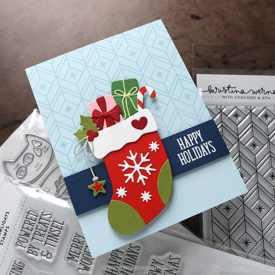 A handmade Christmas greeting card with a red stocking, green gift box, candy cane, holly, and a navy banner reading “HAPPY HOLIDAYS,” photographed on a tabletop with stamp and embosser tools around it.