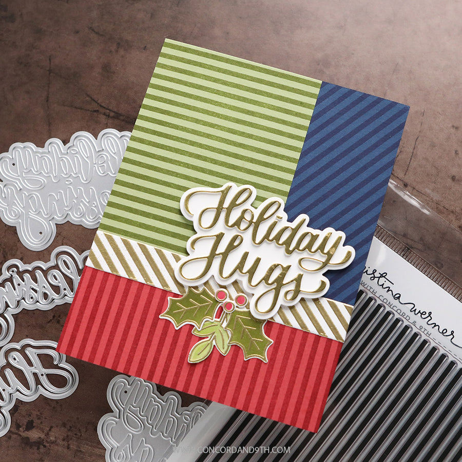 A handmade holiday greeting card with green, navy, and red striped sections, a gold 'Holiday Hugs' script overlay, and a green holly leaf embellishment, photographed on a rustic brown surface with die-cut embellishments nearby.