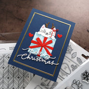 A handmade Christmas greeting card featuring a white cat peeking out of a light blue gift box with a red bow, set on a navy blue background with gold trim. The card has 'Merry Christmas' written in white script and is styled with clear stamps and dies on a brown textured surface.