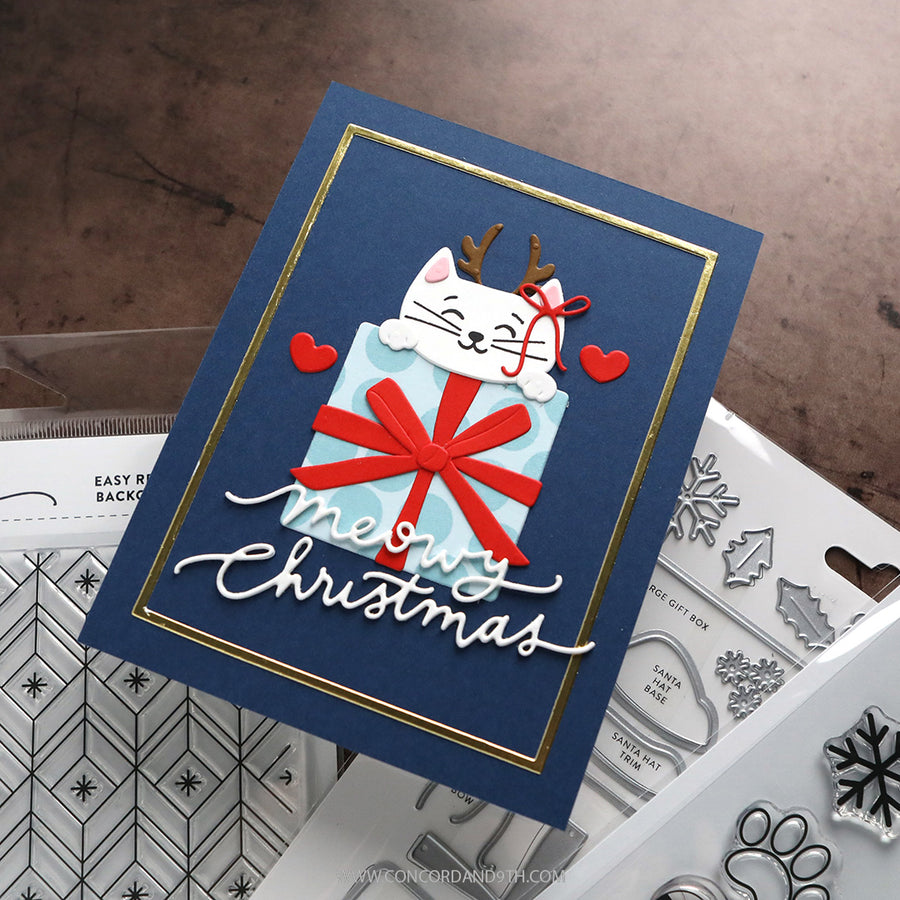A handmade Christmas greeting card featuring a white cat peeking out of a light blue gift box with a red bow, set on a navy blue background with gold trim. The card has 'Merry Christmas' written in white script and is styled with clear stamps and dies on a brown textured surface.