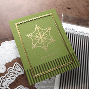 A handmade holiday greeting card with an olive green background, a gold foil snowflake design, and scalloped gold borders, styled on a brown textured surface with crafting dies and a stamp visible.