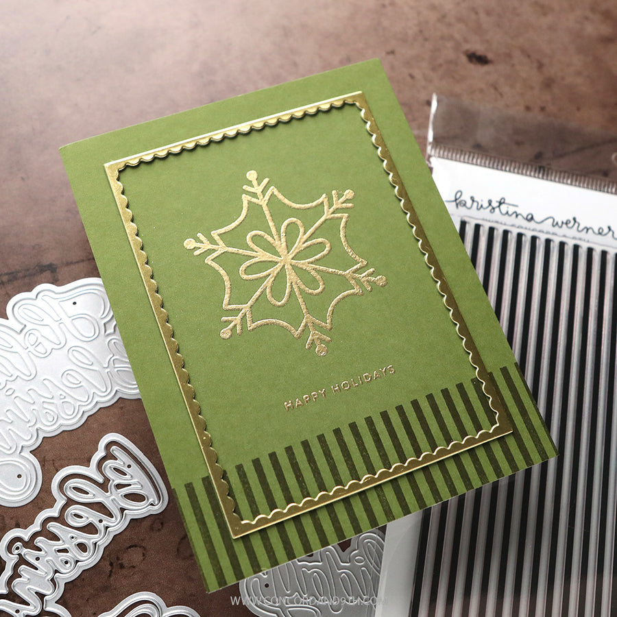 A handmade holiday greeting card with an olive green background, a gold foil snowflake design, and scalloped gold borders, styled on a brown textured surface with crafting dies and a stamp visible.