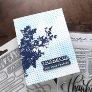 A handmade greeting card featuring a navy blue floral silhouette on a light blue and white halftone background, with the text 'THANKFUL FOR YOUR PRAYERS' in layered black and white. The card is displayed on top of clear stamp blocks and metal dies on a brown textured surface.