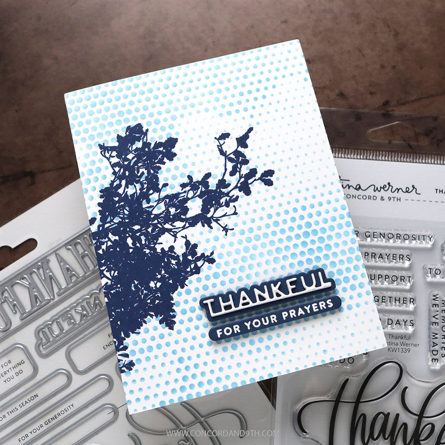 A handmade greeting card featuring a navy blue floral silhouette on a light blue and white halftone background, with the text 'THANKFUL FOR YOUR PRAYERS' in layered black and white. The card is displayed on top of clear stamp blocks and metal dies on a brown textured surface.