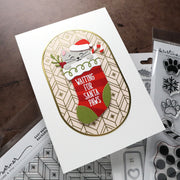 A handmade Christmas greeting card featuring a white cat in a Santa hat peeking out of a red stocking with the text 'WAITING FOR SANTA Paws'. The card is styled on a dark wooden table with crafting tools and clear stamp packages visible in the background.
