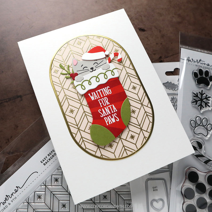 A handmade Christmas greeting card featuring a white cat in a Santa hat peeking out of a red stocking with the text 'WAITING FOR SANTA Paws'. The card is styled on a dark wooden table with crafting tools and clear stamp packages visible in the background.