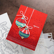 A handmade Christmas greeting card with a textured red background, featuring two layered hot air balloon ornaments in red, green, and pink, tied with white string. The card has the word 'reprise' written on it and is styled with metal dies on a rustic brown background.