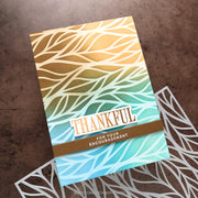 A handmade greeting card with a leaf pattern and a gold-to-blue gradient, featuring the text 'THANKFUL FOR YOUR ENCOURAGEMENT' on a brown band, displayed on a textured brown surface with white laser-cut die-cut accents.