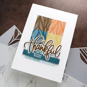 A handmade layered greeting card with a white cardstock base, featuring a colorful geometric pattern in teal, brown, orange, and yellow, with a die-cut script 'thankful' and a small 'BEYOND WORDS' tag. The card is displayed on a textured brown surface with other white cards partially visible underneath.