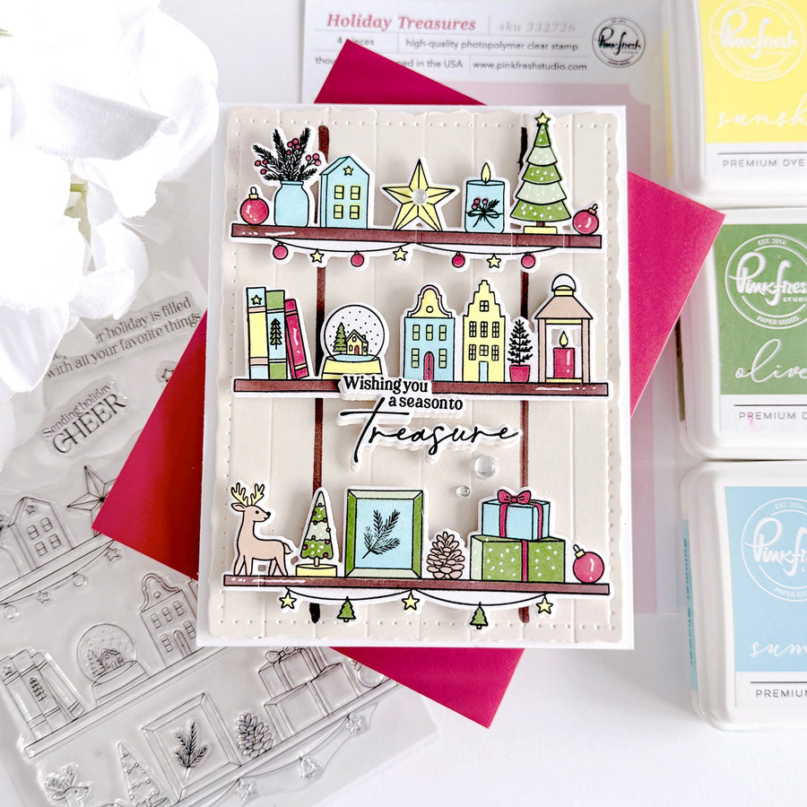 A handmade Christmas-themed card displayed on a white surface with red paper and stamping supplies around it. The card features illustrated holiday shelves with houses, trees, gifts, stars, and the phrase 'Wishing you a season to Treasure.