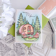 A handmade greeting card featuring an illustrated pink camper trailer, two pink chairs, evergreen trees, and the phrase 'That's how it's done!' in script. The card is styled on a green paper backing with crafting tools and white flowers around it.