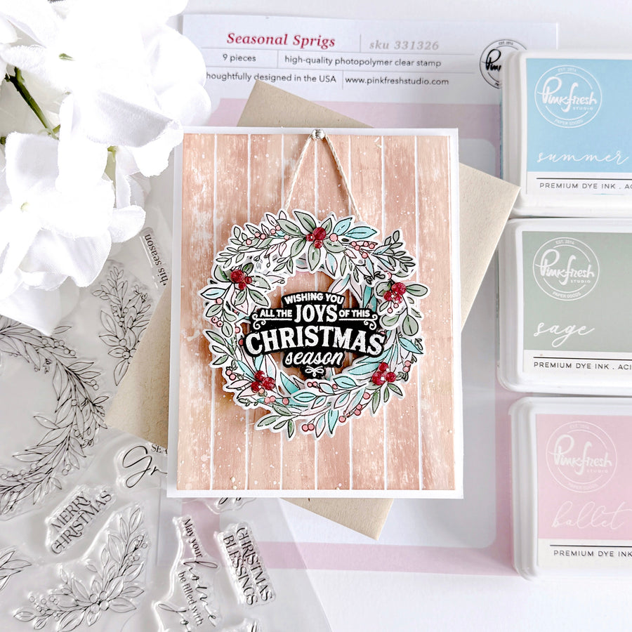A handmade Christmas greeting card with a floral wreath design and the words 'MERRY CHRISTMAS' and 'JOYS OF THE CHRISTMAS season' is displayed on a kraft envelope. The card is styled with a pink striped background, a kraft envelope, white flowers, and a collection of 'Pinwheel' brand stamp and ink pads.