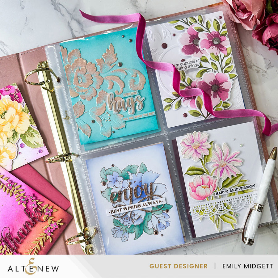 A close-up of a floral guest-designer album page in a clear binder, showing four decorated cards or inserts with pink, teal, blue, and white floral artwork, ribbons, and decorative text. A pen, additional album pages, and flowers are arranged around it on a marble surface.
