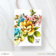 A floral handmade greeting card featuring a colorful watercolor-style bouquet with yellow, pink, blue, and green flowers on a white card, photographed on a light marble surface. The card has the phrase 'share the joy' printed in black script near the lower right.