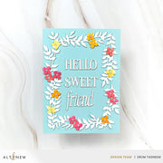 A handmade greeting card with a light blue background, featuring a white die-cut floral border and the words 'HELLO SWEET friend' in white script. The card is decorated with small layered paper flowers in pink, yellow, and orange.