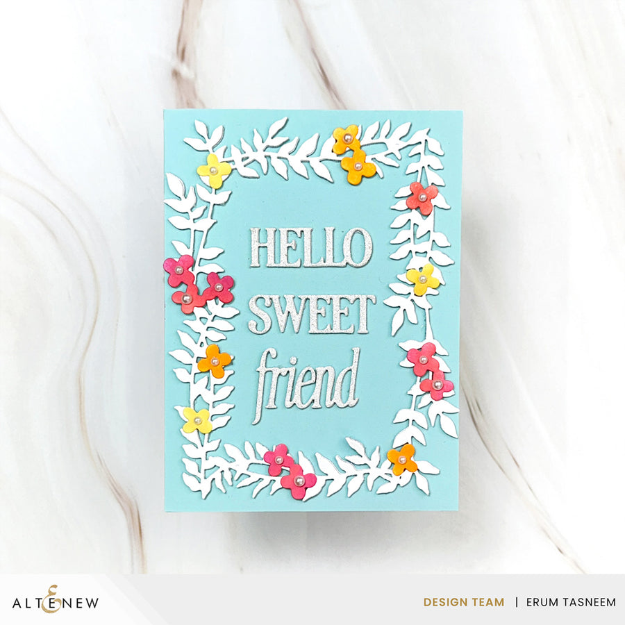 A handmade greeting card with a light blue background, featuring a white die-cut floral border and the words 'HELLO SWEET friend' in white script. The card is decorated with small layered paper flowers in pink, yellow, and orange.