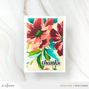 A vertical handmade greeting card with a white border, featuring a colorful floral design in coral, burgundy, teal, and green on a cream background, with the word 'thanks' in black script near the bottom. The card is photographed on a light marble surface, with Altenew branding and designer credits at the bottom of the frame.