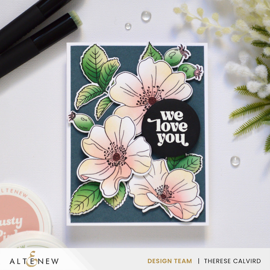 A handmade greeting card featuring layered white and peach flowers with green leaves on a dark teal background, with a black circular sticker reading 'we love you'. The card is photographed on a white surface with craft supplies and small flowers around it. Altenew One-Go: Briar Blossoms card ideas