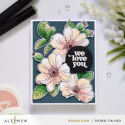 A handmade greeting card featuring layered white and peach flowers with green leaves on a dark teal background, with a black circular sticker reading 'we love you'. The card is photographed on a white surface with craft supplies and small flowers around it. Altenew One-Go: Briar Blossoms card ideas