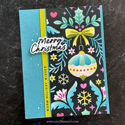 A handmade Christmas greeting card featuring a turquoise blue left panel, a vertical yellow stripe, and a black right panel decorated with teal holly leaves, pink hearts, yellow stars, a yellow bow, and a pastel ornament. A die-cut sticker reads “Merry Christmas,” and the card is photographed on a dark textured surface.