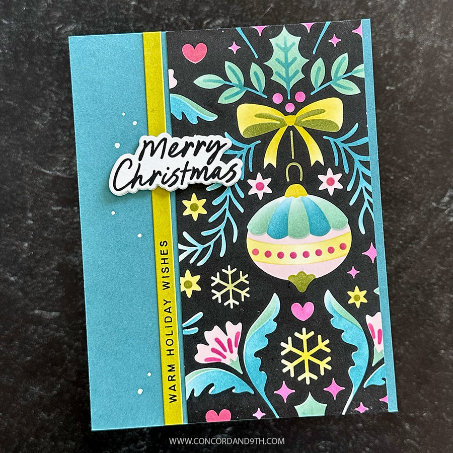 A handmade Christmas greeting card featuring a turquoise blue left panel, a vertical yellow stripe, and a black right panel decorated with teal holly leaves, pink hearts, yellow stars, a yellow bow, and a pastel ornament. A die-cut sticker reads “Merry Christmas,” and the card is photographed on a dark textured surface.