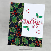 A handmade Christmas greeting card featuring a white center with the word 'merry' in red script, green holly leaves, and red berries, layered over a dark navy and green patterned holly background. The card is shown at an angle on a neutral gray background.