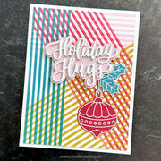 A handmade holiday greeting card with layered die-cut elements, featuring the words 'Holiday Hugs' in white script over a background of pink, teal, yellow, and red striped and checkered patterns. A red ornament and teal holly leaf are attached at the bottom.