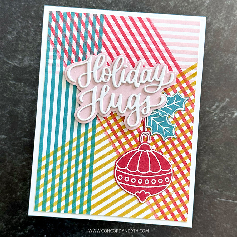 A handmade holiday greeting card with layered die-cut elements, featuring the words 'Holiday Hugs' in white script over a background of pink, teal, yellow, and red striped and checkered patterns. A red ornament and teal holly leaf are attached at the bottom.