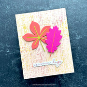 A handmade greeting card featuring two layered autumn leaves, one coral-pink and one bright pink, with yellow and green paper veins. The card has a textured cream background with subtle red and yellow speckles and a die-cut sticker reading 'HEARTFELT hugs' at the bottom.