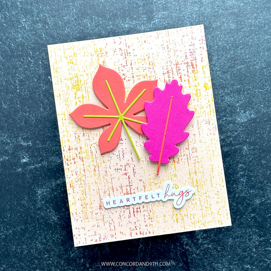 A handmade greeting card featuring two layered autumn leaves, one coral-pink and one bright pink, with yellow and green paper veins. The card has a textured cream background with subtle red and yellow speckles and a die-cut sticker reading 'HEARTFELT hugs' at the bottom.